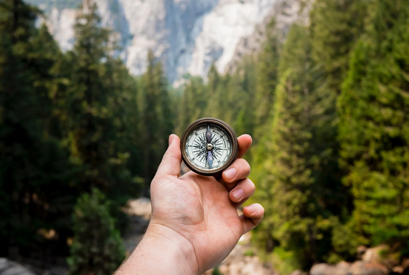 Close-up of a hand holding a compass against a backdrop of green forest and distant mountains, natural adventurous mood